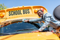 smiling mature african american bus driver wiping front window of school bus and looking at camera